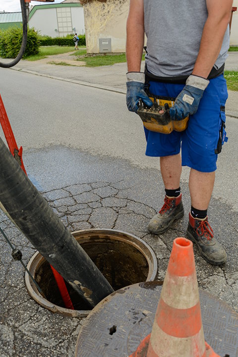 Arbeiter bei Kanalwartung neben offenem Gully und Warnkegel.